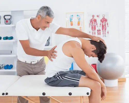 A man is sitting on the table while another person stands near him.